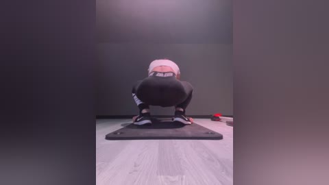 Media: Video of a person squatting with a white headband, black leggings, and black shoes, on a yoga mat in a dimly lit room with light wooden flooring.