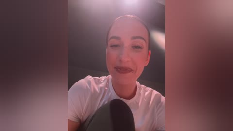 Media: A video of a woman with fair skin, dark hair, and light makeup, wearing a white t-shirt, speaking into a microphone in a dimly lit room with blurred background.