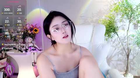 Media: A video of an Asian woman with fair skin, black hair, and glasses, sitting on a white chair in a room with a floral arrangement and a potted plant.