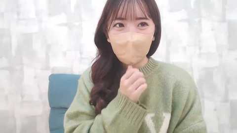 Media: A video of a young Asian woman with long, dark hair, wearing a beige surgical mask, a green sweater, and a blue chair, indoors with white curtains.