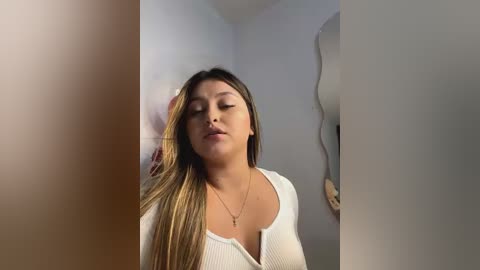 Media: Video of a Latina woman with long, straight, blonde hair, wearing a white ribbed sweater, standing in a minimalist room with white walls and a large, abstract mirror.