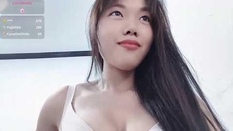 Media: Video of a young Asian woman with long black hair, fair skin, and a slender physique, wearing a white bra. Background shows a digital screen with live stats and a blurred white wall.