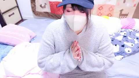 Media: Video of a young Asian woman with straight black hair, wearing a blue cap, light gray sweater, and white face mask, kneeling on a bed with pastel-colored pillows and a stuffed animal.