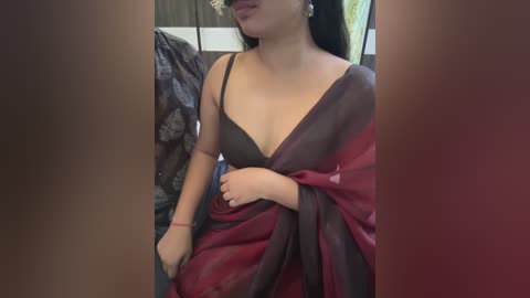 Media: Video of an Indian woman with medium skin tone and long black hair, wearing a black bra and a red and black sari, partially draped, sitting indoors with blurred background.