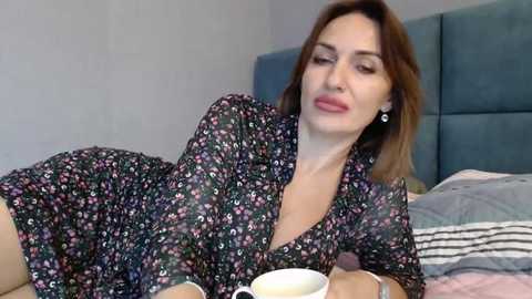 Media: Video of a Caucasian woman with light skin, shoulder-length brown hair, wearing a black floral dress, lying on a bed with a blue headboard, holding a cup, looking relaxed.