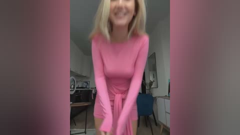 Media: Video of a smiling blonde woman in a pink dress, standing in a modern kitchen with white cabinets and a blue chair.