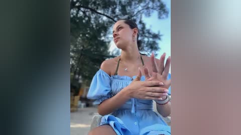 Media: Video of a fair-skinned woman with brunette hair in a light blue off-shoulder dress, seated outdoors under a tree. She gestures with her hands. Background shows blurred greenery and sky.