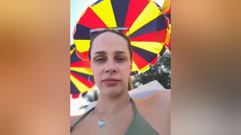 Media: Video of a woman with fair skin, dark hair, and a medium build, wearing a green bikini, holding a vibrant, multicolored umbrella with yellow and red stripes. Background features blurred trees and sky.