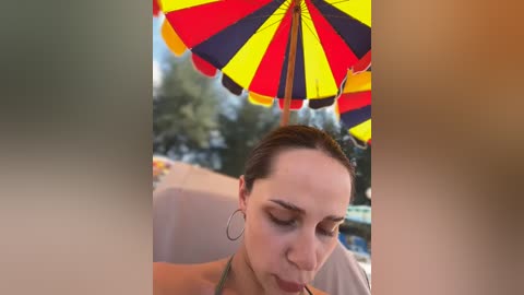 Media: Video of a woman with light skin and brown hair, wearing a green bikini, eating a popsicle under a multi-colored umbrella. Background features blurred greenery.