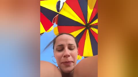 Media: Video of a fair-skinned woman with closed eyes, under a vibrant, multi-colored umbrella with red, yellow, and blue panels against a clear blue sky, capturing a relaxed and sunny outdoor scene.