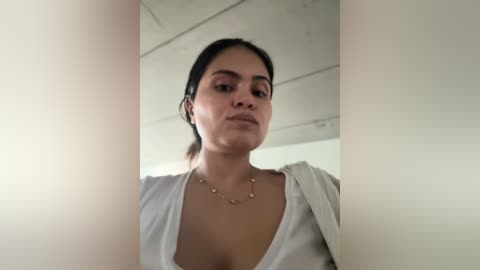 Media: A low-angle video of a Latina woman with dark hair, wearing a white blouse, necklace, and headphones, standing in a modern, minimalist room with white walls and a ceiling.