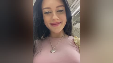 Media: Video of a young woman with long, straight black hair, fair skin, and pink lipstick, wearing a pink top and a heart-shaped necklace, smiling slightly.