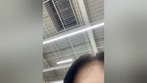 Media: A video of a person with short dark hair, taken from a low angle inside a high-ceilinged, industrial-style room with exposed beams, white ceiling panels, and fluorescent lights. The focus is on the ceiling and the person's upper body.