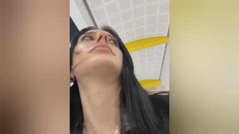 Media: Video of a woman with long black hair and olive skin, wearing a black top, lying back with a slight smile, in a room with white grid ceiling and yellow accents.