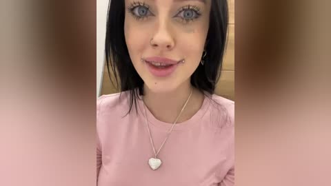 Media: Video of a young woman with fair skin, blue eyes, and black hair, wearing a pink shirt and a silver heart necklace. She has a nose piercing and a lip piercing. Background is blurred.