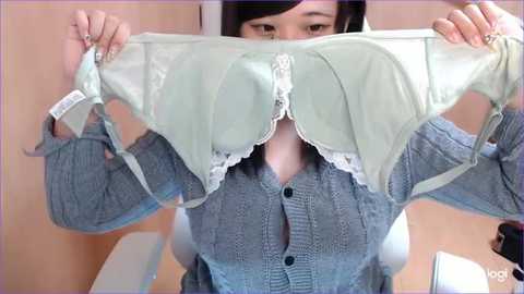 Media: A video of an Asian woman with dark hair, wearing a grey cardigan, holding up a mint green bra with white lace trim, against a light beige wall.