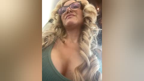 Media: Video of a blonde woman with curly hair, wearing glasses, a low-cut teal top, and a confident expression.