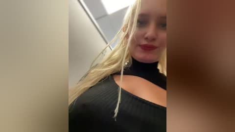 Media: A video of a blonde woman with a fair complexion, wearing a black, ribbed turtleneck, smiling, taken from a low angle in a dimly lit, narrow hallway.