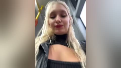 Media: Video of a blonde woman with long hair, wearing a black leather jacket and a black top, sitting on a subway.