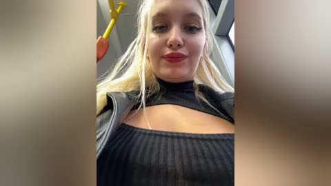 Media: Video of a blonde woman with fair skin, wearing a black ribbed top and a black jacket, making a duck face in a public space with yellow handrails.