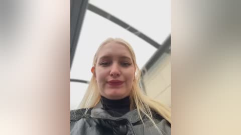 Media: A close-up video of a young, smiling Caucasian woman with long, straight blonde hair, wearing a black jacket, standing in a brightly lit, modern building with large glass windows.