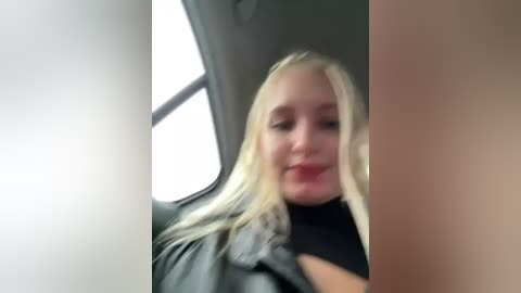 Media: A blurry video shows a smiling blonde woman in a black turtleneck sweater and jacket, seated in a car with light-colored interior.