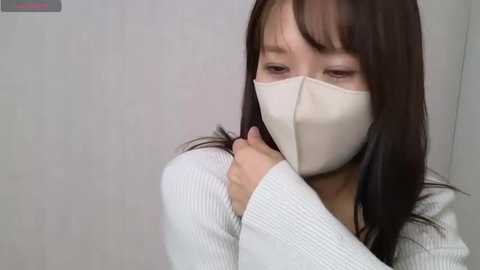 Media: Video of an East Asian woman with long, straight black hair and light skin, wearing a white face mask and a ribbed white sweater. She is holding her right hand to her mouth, looking concerned. The background is a plain, light-colored wall.