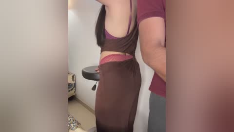 Media: Video of a light-skinned woman in a dark brown tank top and matching sheer shorts, showing her midriff and buttocks, standing in a small, beige-tiled bathroom with a floral rug.