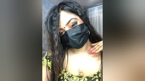 Media: Video of a young woman with long, wavy black hair, wearing a black face mask and a floral-patterned yellow top, with a necklace featuring a pendant. She has medium skin tone and is indoors, with a blurred background.