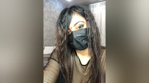 Media: Video of a young Asian woman with long, wavy black hair, wearing a black fishnet top, face mask, and dark makeup. She has a medium skin tone. Background shows a gray wall and white curtains.