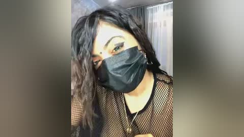 Media: Video of a person with long, wavy black hair, wearing a black fishnet top, black face mask, and a silver necklace. Background shows a blurred indoor setting with a white curtain.