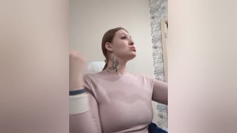 Media: Video of a Caucasian woman with fair skin, light brown hair, and a tattoo on her neck, wearing a pink top, standing in a bathroom with a tiled wall and white sink.