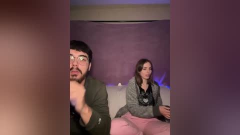 Media: Video of two people watching a screen in a dimly lit room. Man with glasses and beard, woman with long brown hair, wearing a grey jacket.