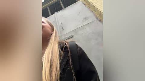 Media: Video of a blonde woman with long hair, wearing a black jacket, walking on a grey concrete surface. The background includes a yellow railing and a partially visible grey wall.