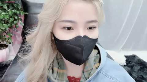 Media: A video of an East Asian woman with long, blonde hair and fair skin, wearing a black face mask, denim jacket, and a green plaid shirt, sitting indoors with a potted plant and a black fur rug in the background.