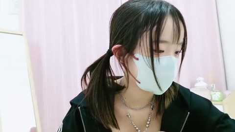 Media: Video of an Asian woman with long black hair in pigtails, wearing a white face mask, black jacket, and silver necklace, indoors with light pink curtains and a shelf with bottles in the background.