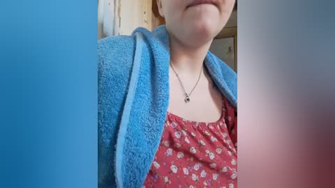 Media: A close-up video of a fair-skinned woman with light brown hair, wearing a red floral top and a blue towel over her shoulders, with a silver necklace. The background is blurry, possibly a bathroom.