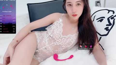 Media: Video of a fair-skinned, slender, young woman with long dark hair, wearing a sheer, white lace bodysuit, lying on a white bed with black pillows and a pink vibrator. Background shows a digital interface with heart icons.