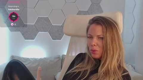 Media: A video of a light-skinned woman with long, blonde hair, wearing a black robe, reclining on a white leather chair in a modern, minimalist room with hexagonal grey wall panels.