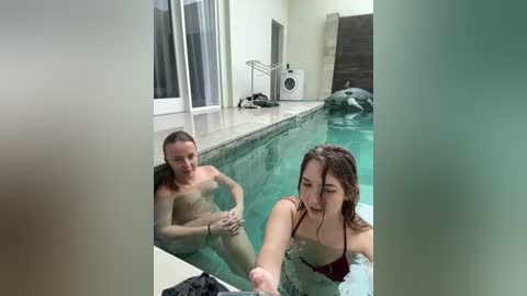 Media: Video of a man and woman in a pool, both shirtless, with a dolphin swimming nearby. The indoor pool is clean, with a tiled edge and white walls.