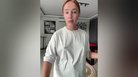 Media: A video of a young Caucasian woman with fair skin and light brown hair, wearing a loose white t-shirt, standing in a modern, minimalist room with white walls, a black sofa, and abstract artwork.