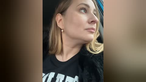 Media: Video of a young Caucasian woman with shoulder-length blonde hair, wearing a black t-shirt with a white design, and a fur coat, looking upward in a car.