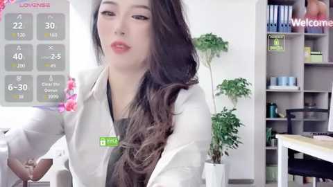 Media: Video of an Asian woman with long, wavy brown hair, wearing a white lab coat, sitting in an office with a plant and a bookshelf in the background.