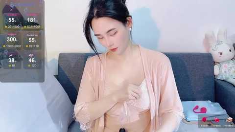 Media: A video of a young Asian woman with fair skin, dark hair, wearing a pink robe, adjusting her white lace bra, in a cozy living room with a gray couch, stuffed toys, and a digital health tracker overlay.