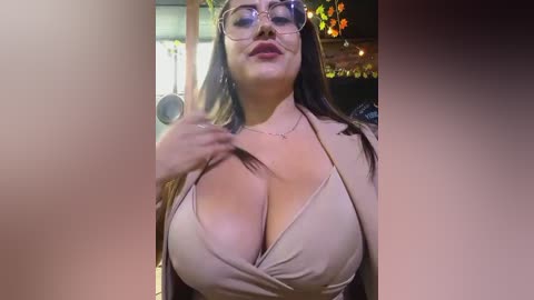 Media: Video of a plus-sized woman with long brown hair and glasses, wearing a beige halter top that reveals ample cleavage, indoors with a blurred background of string lights.