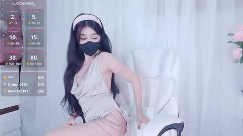 Media: Video of an Asian woman with long black hair, wearing a black face mask, beige dress, and white headband, sitting in a white chair against white curtains.