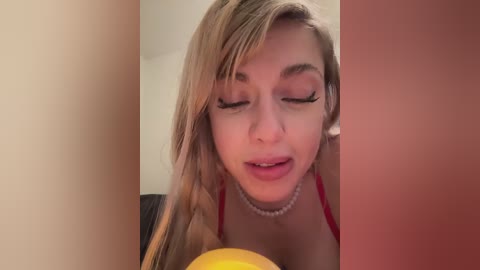 Media: Video of a blonde woman with long hair, wearing a red bra and pearl necklace, holding a yellow object, smiling, in a dimly lit room.