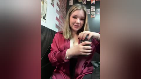 Media: Video of a young woman with straight, shoulder-length blonde hair, wearing a red satin jacket, sitting on a black cushioned bench in a dimly lit, brick-walled room with framed photos on the wall.