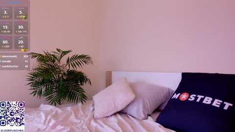 Media: Video of a minimalist bedroom with a white bed, grey pillows, a large potted plant, and a \"MOSTBET\" pillow, set against a beige wall with a digital clock displaying 5:30.