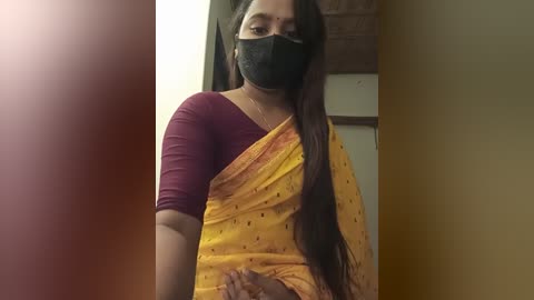 Media: Video of a South Asian woman with long black hair, wearing a maroon top, yellow saree with black polka dots, black face mask, and standing indoors near a door.